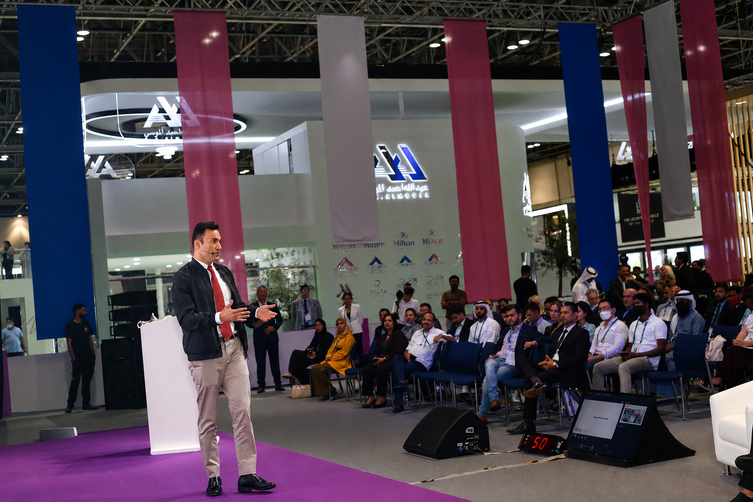 Aladdin Ismail speaking solo on stage beside a podium, seated audience to the right
