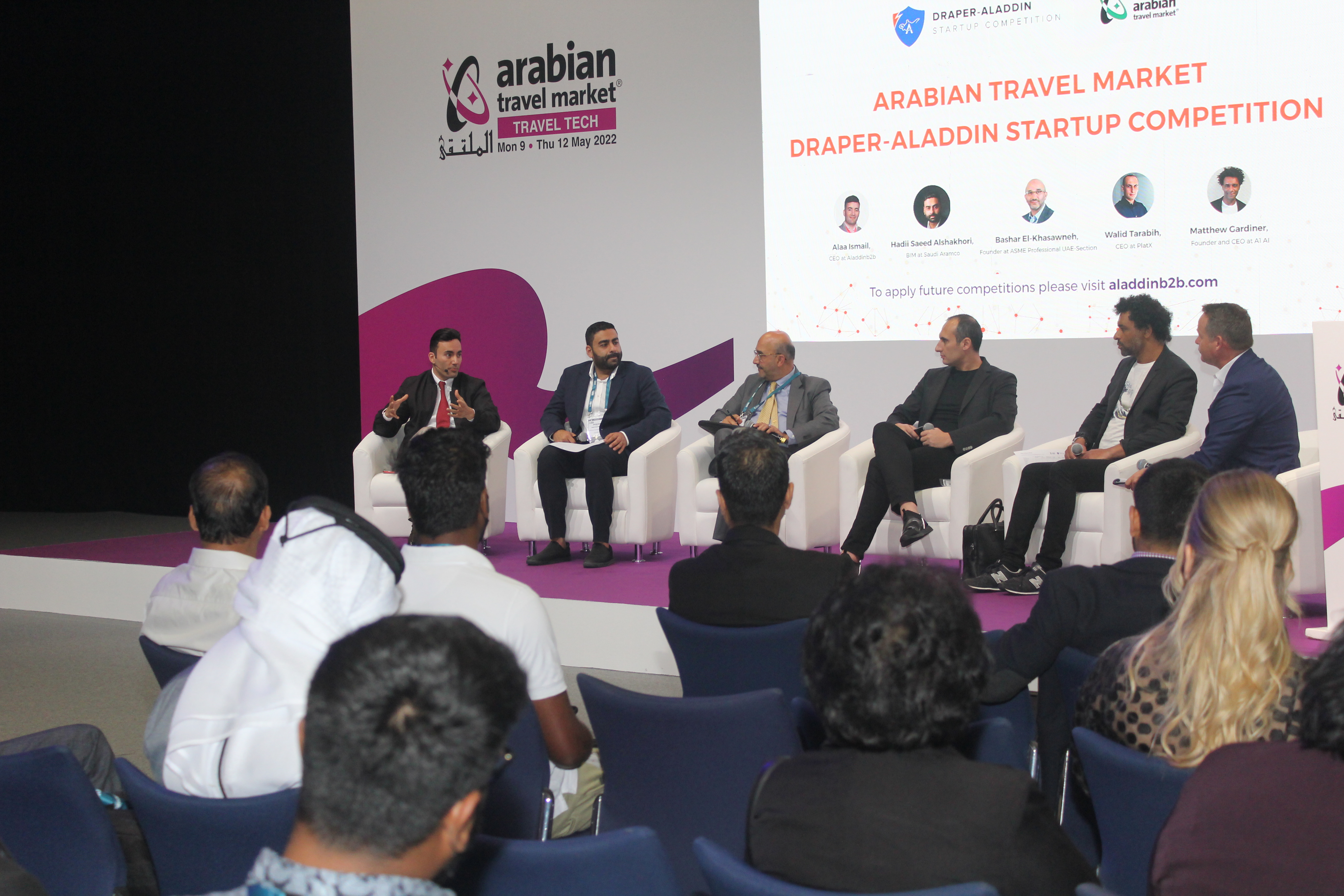 Aladdin Ismail speaking on a panel at Arabian Travel Market, mid gesture, branded backdrop behind the panel
